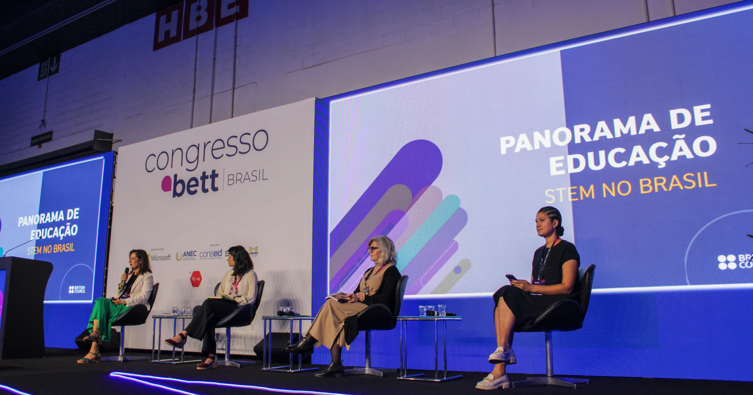 Fotografia do palco do Bett Brasil em que estão presentes Alessandra Moura, Julia Rubiano de La Cruz, Sandra Umbehaum e Ana Paula Gaspar. No telão aparece o texto Panorama de Educação STEM no Brasil